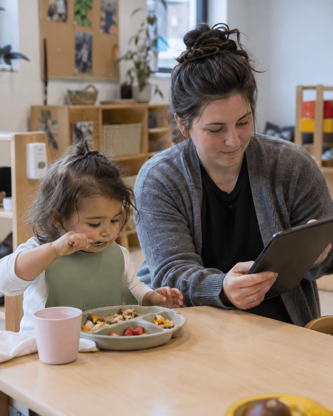 An educator recording a toddler's lunch on a tablet at a small wooden table
